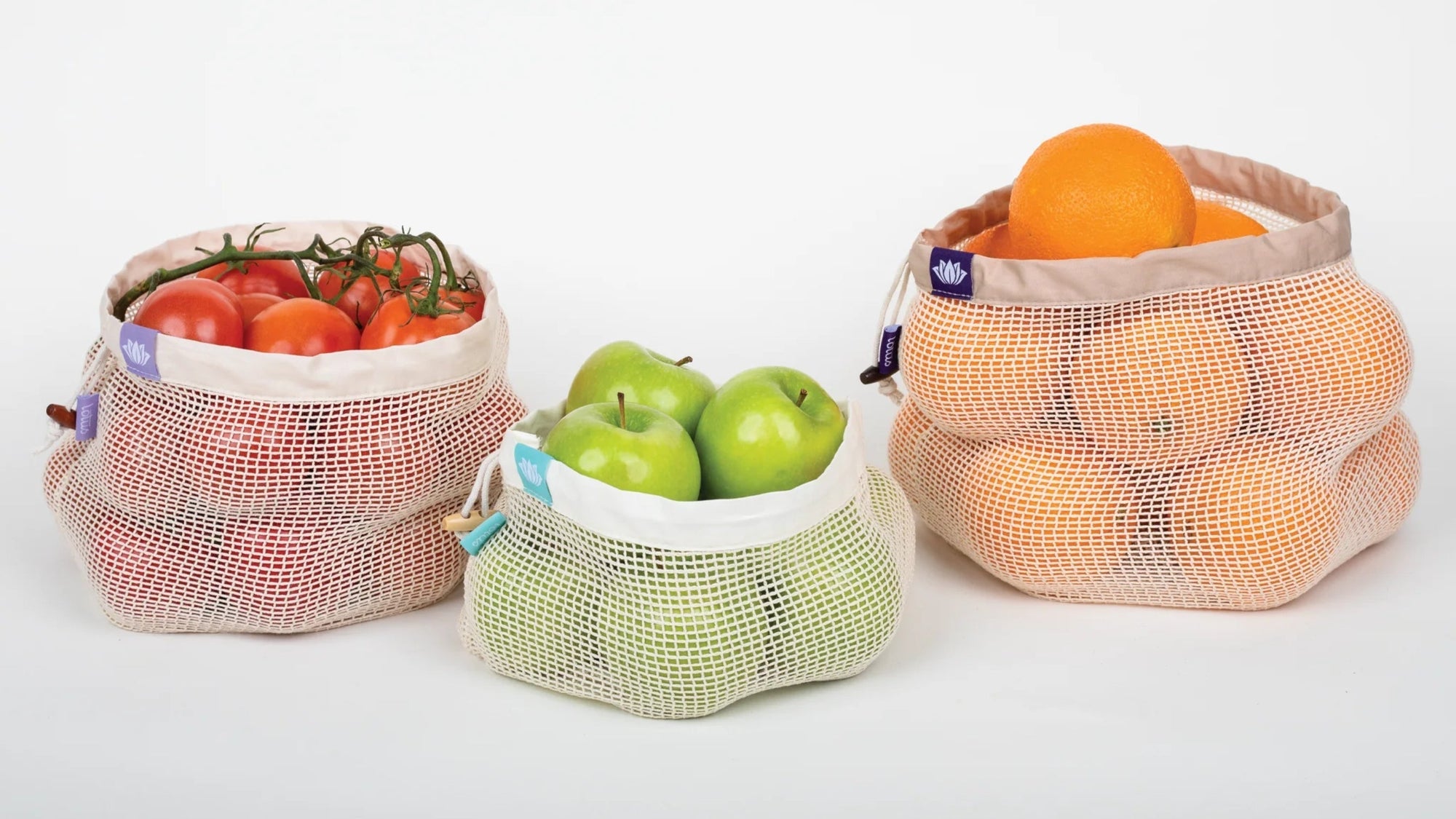 Photo of set of 3 100% Organic Lotus Produce Bags filled with produce. From left to right, medium bag with tomatoes, small bag with green apples, large bag with oranges.